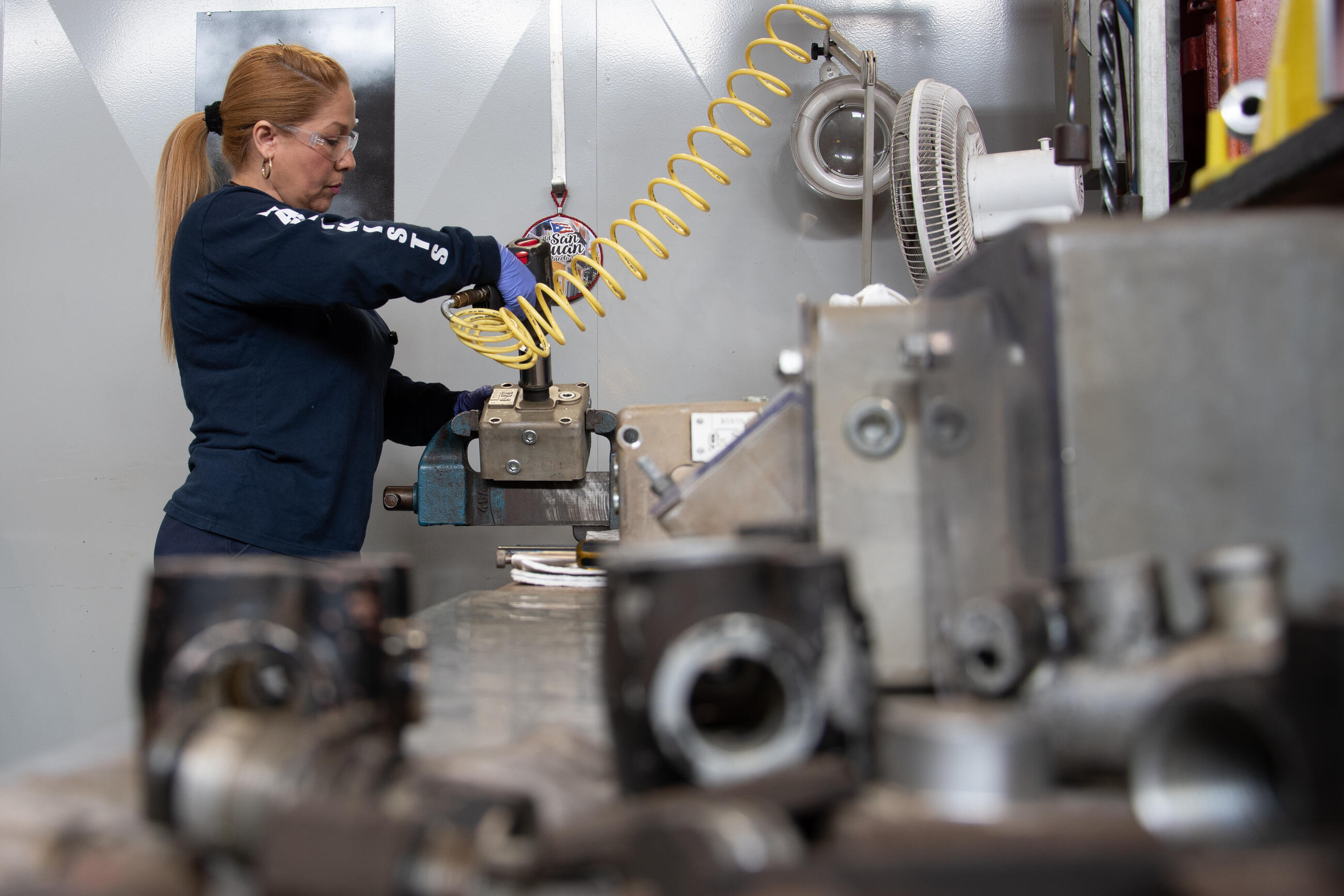 A woman wearing safety glasses uses a power tool on a piece of machinery at a work bench surrounded by steel parts 