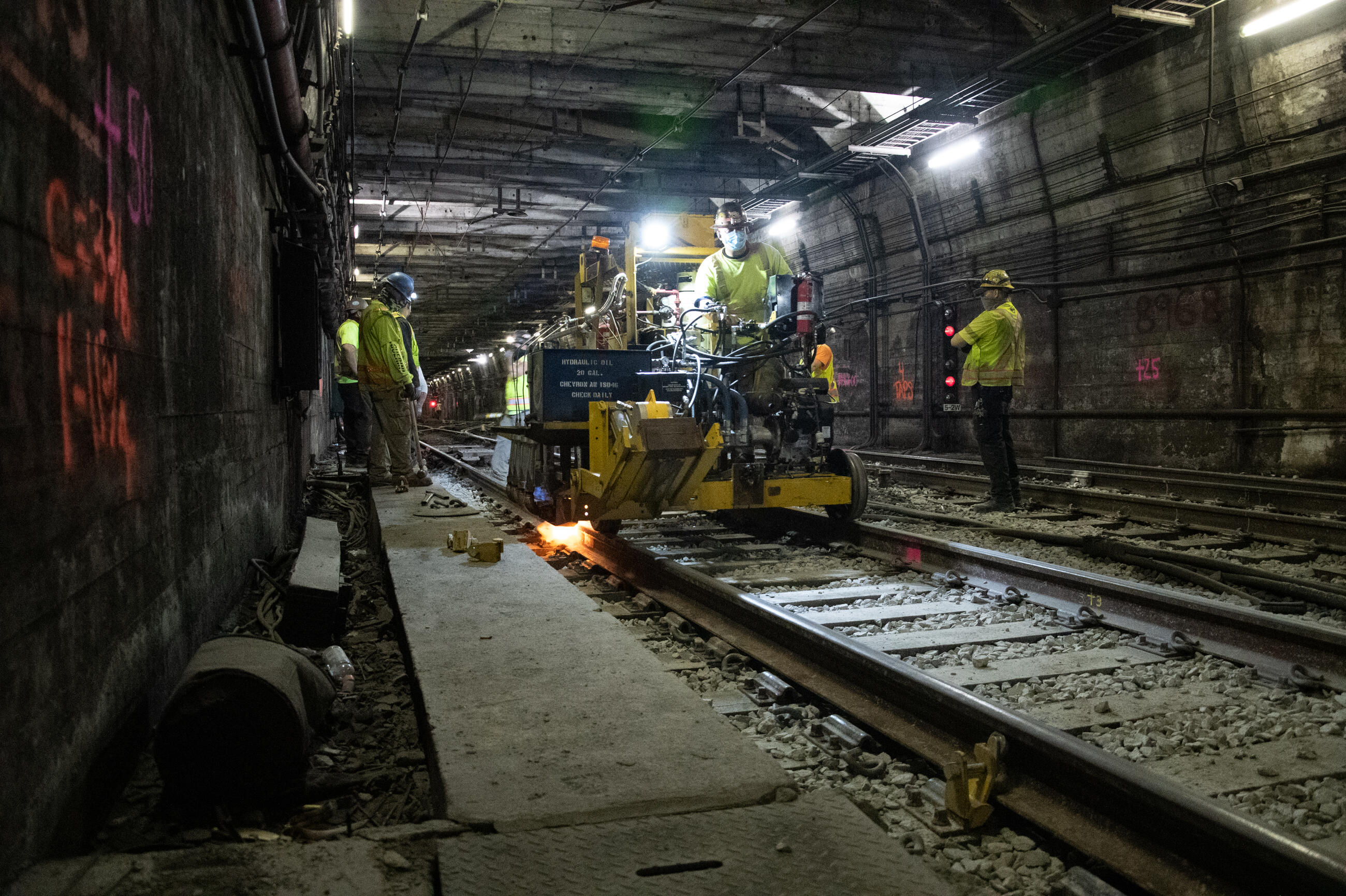 A construction worker drives a piece of machinery over train tracks in a tunnel on the Blue Line. Other workers to the left and the right of the tracks watch as the machinery drives by.