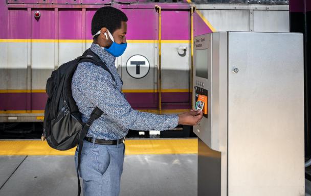 A Commuter Rail rider wearing a face covering uses his CharlieCard at a fare validator at Blue Hill Ave Station.